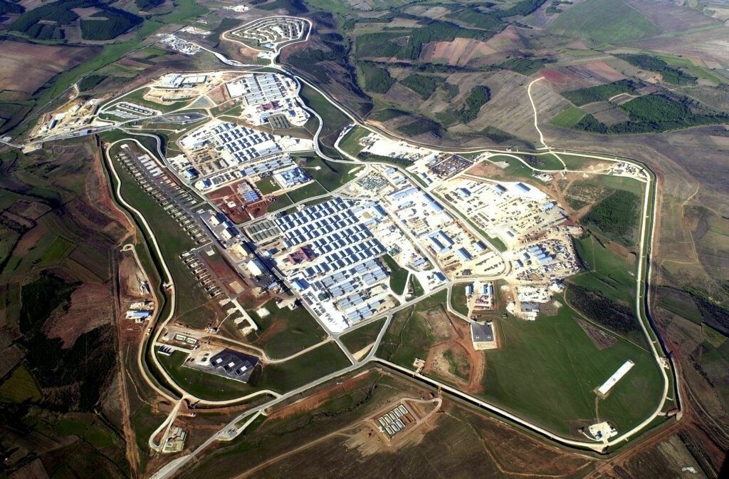 Aerial view of Camp Bondsteel in Kosovo, showing rows of buildings, roads, and surrounding fields.